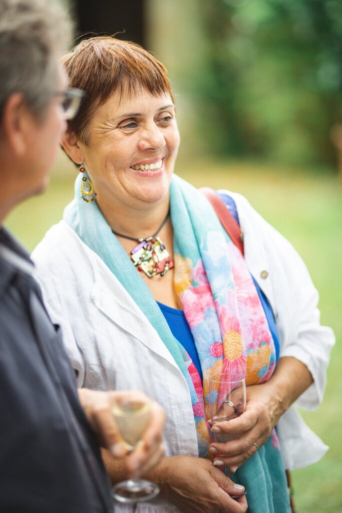Une femme souriante portant un foulard coloré, un verre à la main, en extérieur.