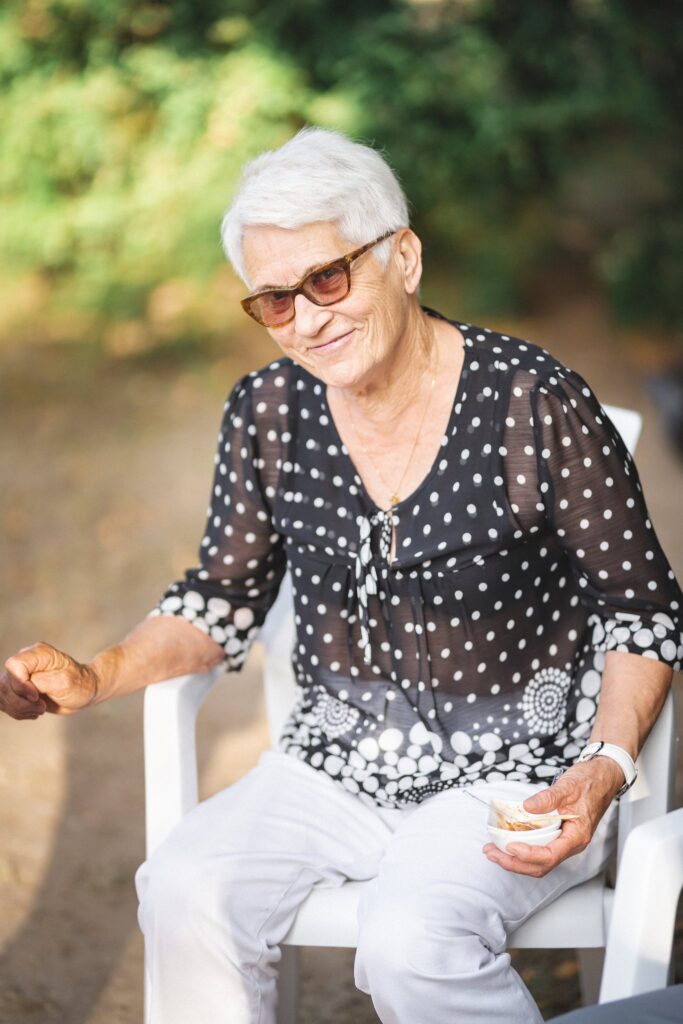 Une femme âgée en lunettes de soleil et chemisier à pois, assise sur une chaise blanche.