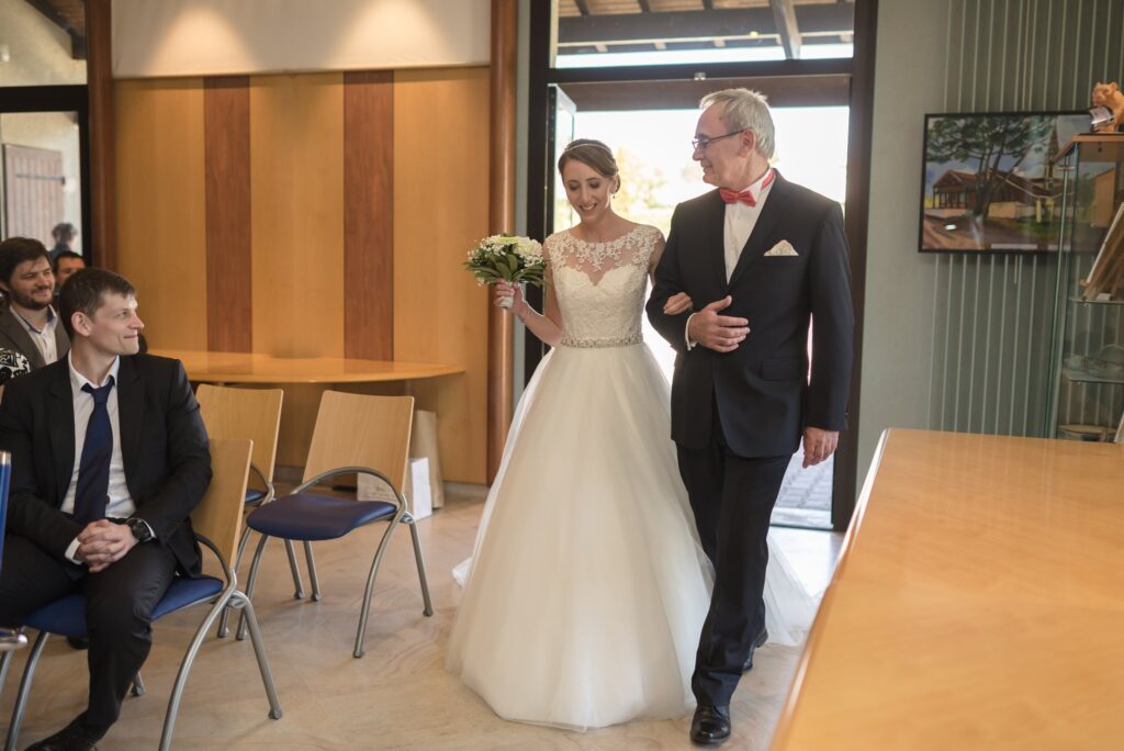 La mariée en robe blanche entre dans la salle de la mairie au bras d'un homme en nœud papillon rouge, devant des invités assis.