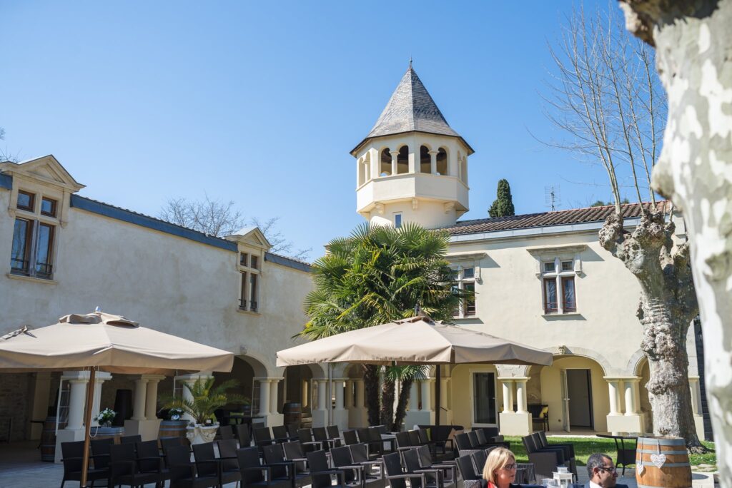 La façade du domaine avec sa tour, des parasols et des rangées de chaises sous un ciel bleu.