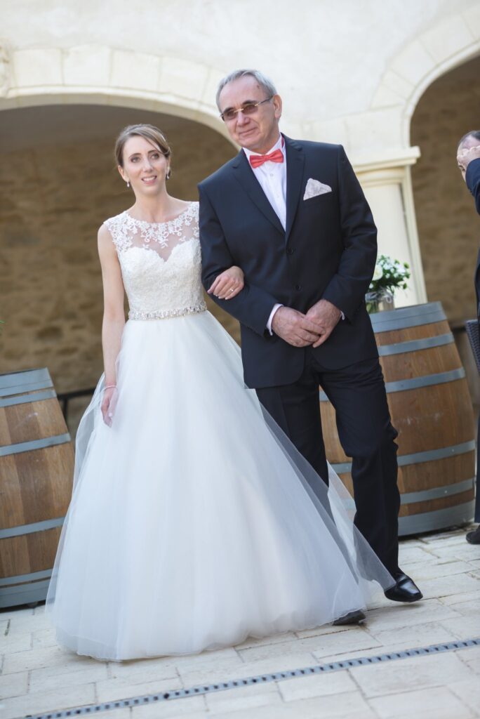 La mariée avance entre des tonneaux au bras d'un homme en costume et nœud papillon rouge.