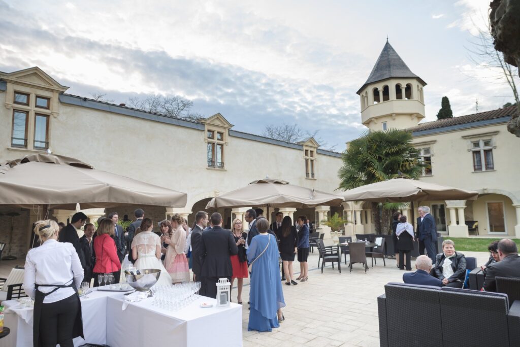 La terrasse du domaine pendant le cocktail, des invités debout sous les parasols et la tour sous un ciel nuageux.