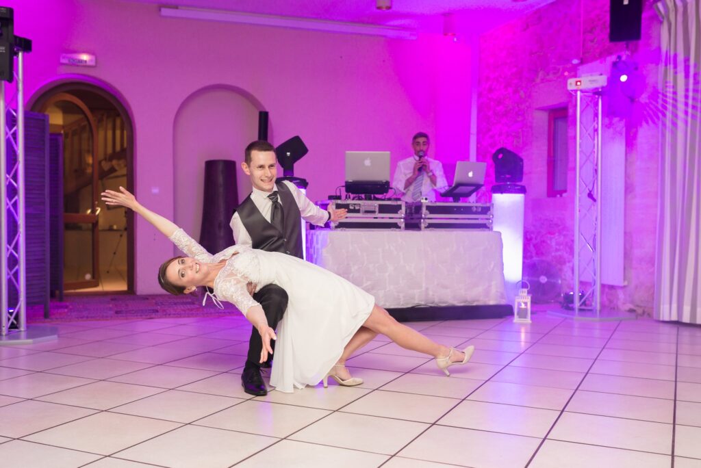 La mariée en robe courte se penche en arrière dans les bras du marié sur la piste de danse éclairée en rose.